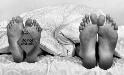 Une femme et un homme couchés au lit, les pieds sortent du drap, une petite pancarte pend sur les pieds de la femme : ne pas déranger (asexalité).
